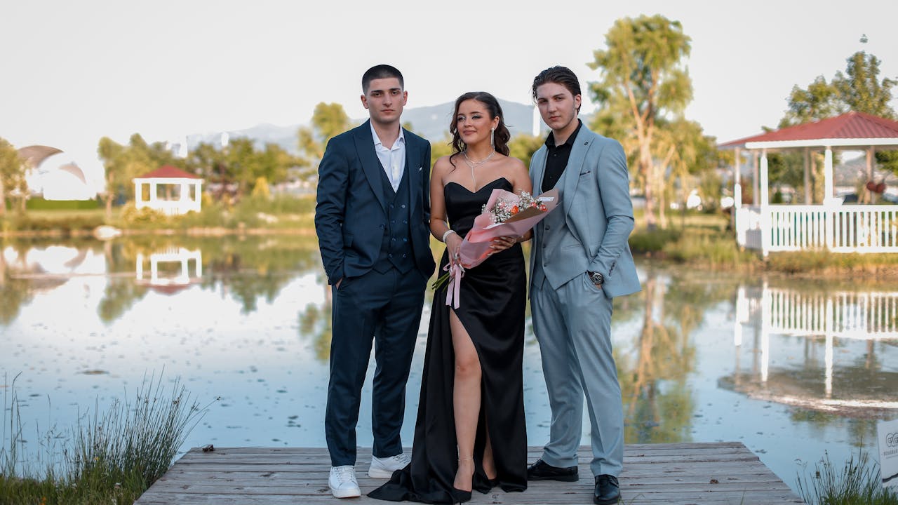 Three elegantly dressed adults pose by a peaceful lakeshore, perfect for fashion or event themes.