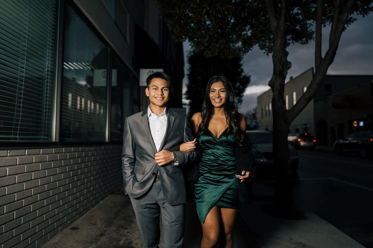A stylish couple walking arm in arm on a city sidewalk at night, dressed in formal attire.